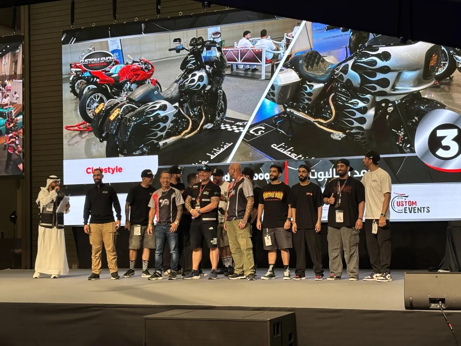 Clubstyle Dubai team on stage at a custom-bike show, their flame-painted bagger on the screen behind