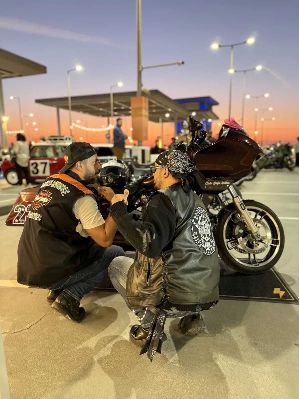 Two riders in club vests by a custom Road Glide at a fuel-station meet at sunset