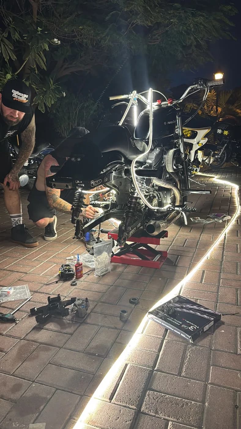 A builder wrenching on a custom Harley on a scissor jack at a late-night driveway meet, LED strip lighting the brick